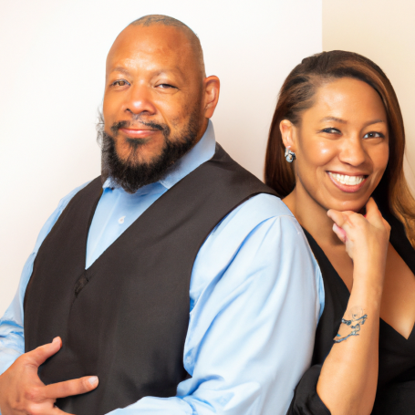 A man and woman smile, dressed formally, posing together against a light background.