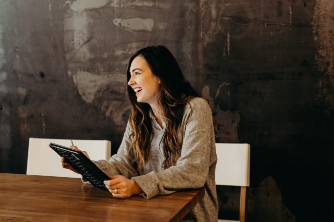 Smiling woman holding a tablet while sitting at a wooden table.